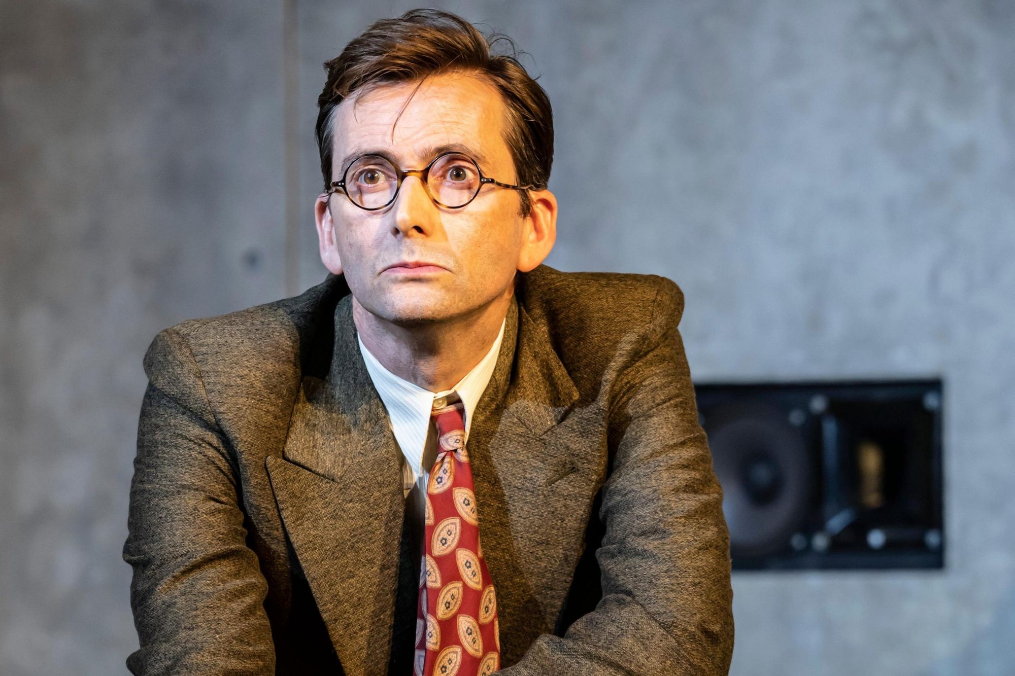 David Tennant as John Halder in a tweed suit and 1930 spectacles, he has a flat expression with a touch of concern