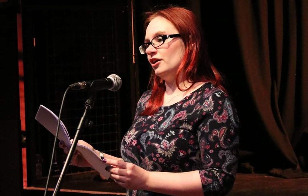 A white woman with long red hair and glasses stands and speaks in front of a microphone holding some paper